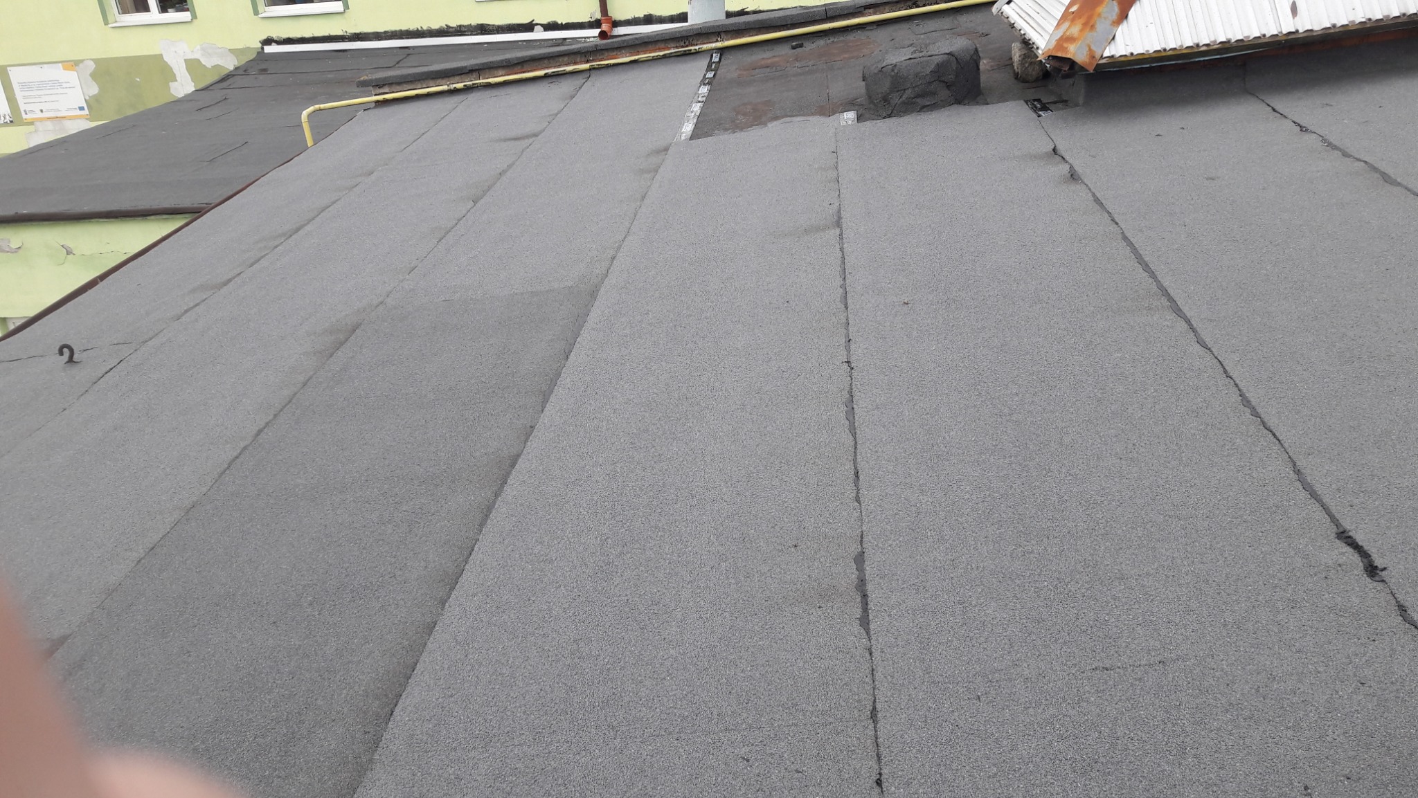 Płaski dach pokryty papą termozgrzewalną, widoczne łączenia pasów, fragment blaszanej obróbki i komin wentylacyjny.