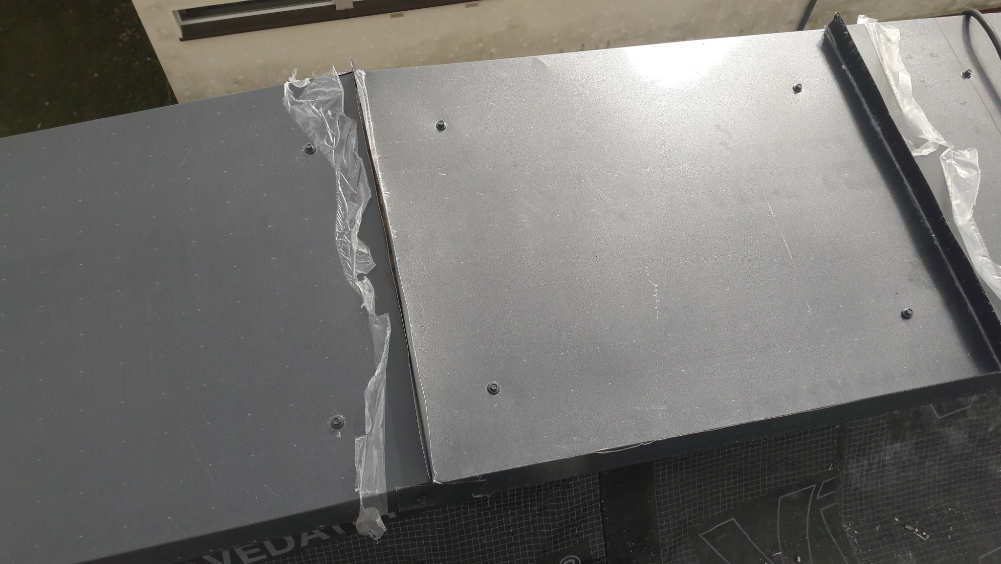 Fragment dachu pokryty szarymi panelami blacharskimi, zabezpieczonymi folią ochronną, widoczne śruby mocujące i fragment membrany dachowej z logotypem.