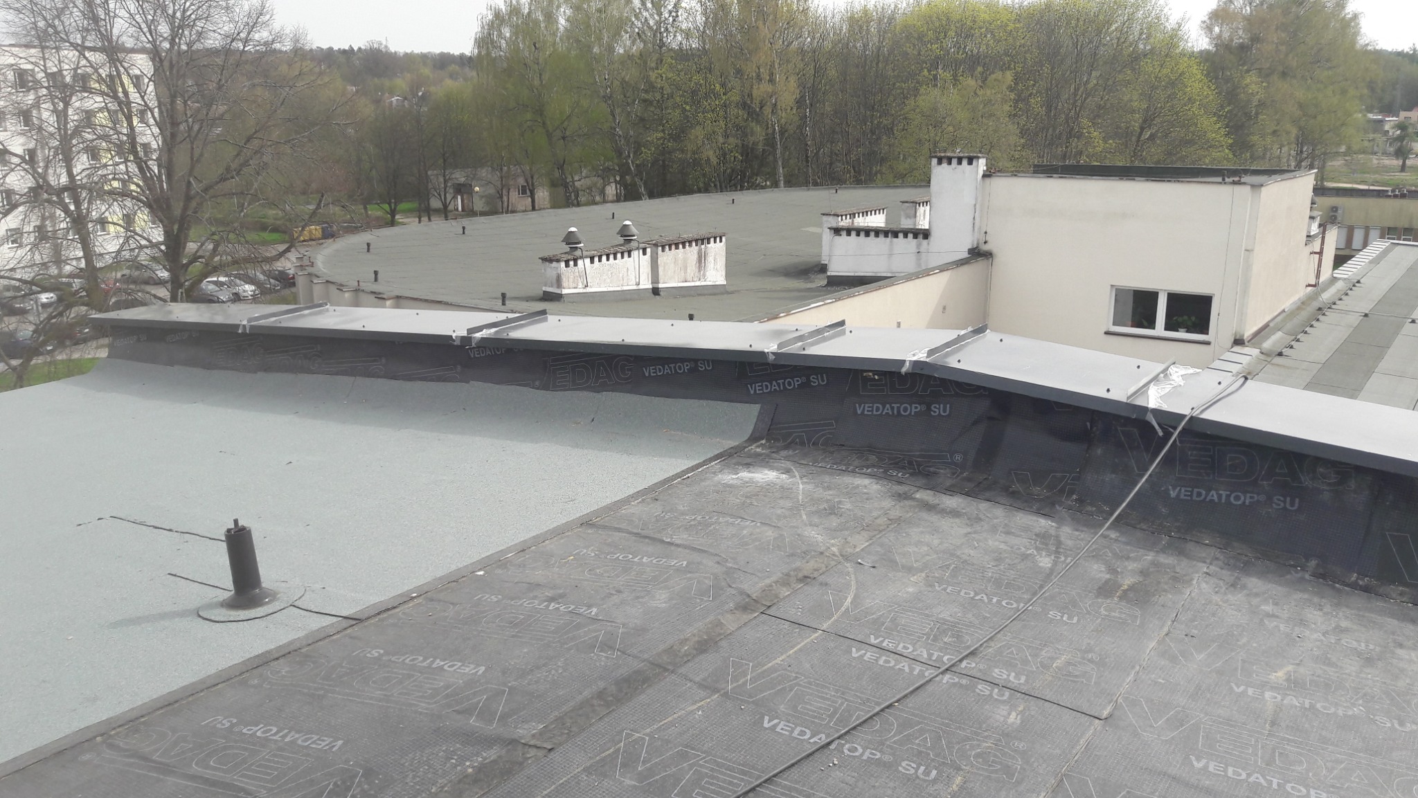 Widok z góry na płaski dach budynku pokryty papą termozgrzewalną z widocznymi łączeniami i logo producenta, w tle budynki mieszkalne i drzewa.