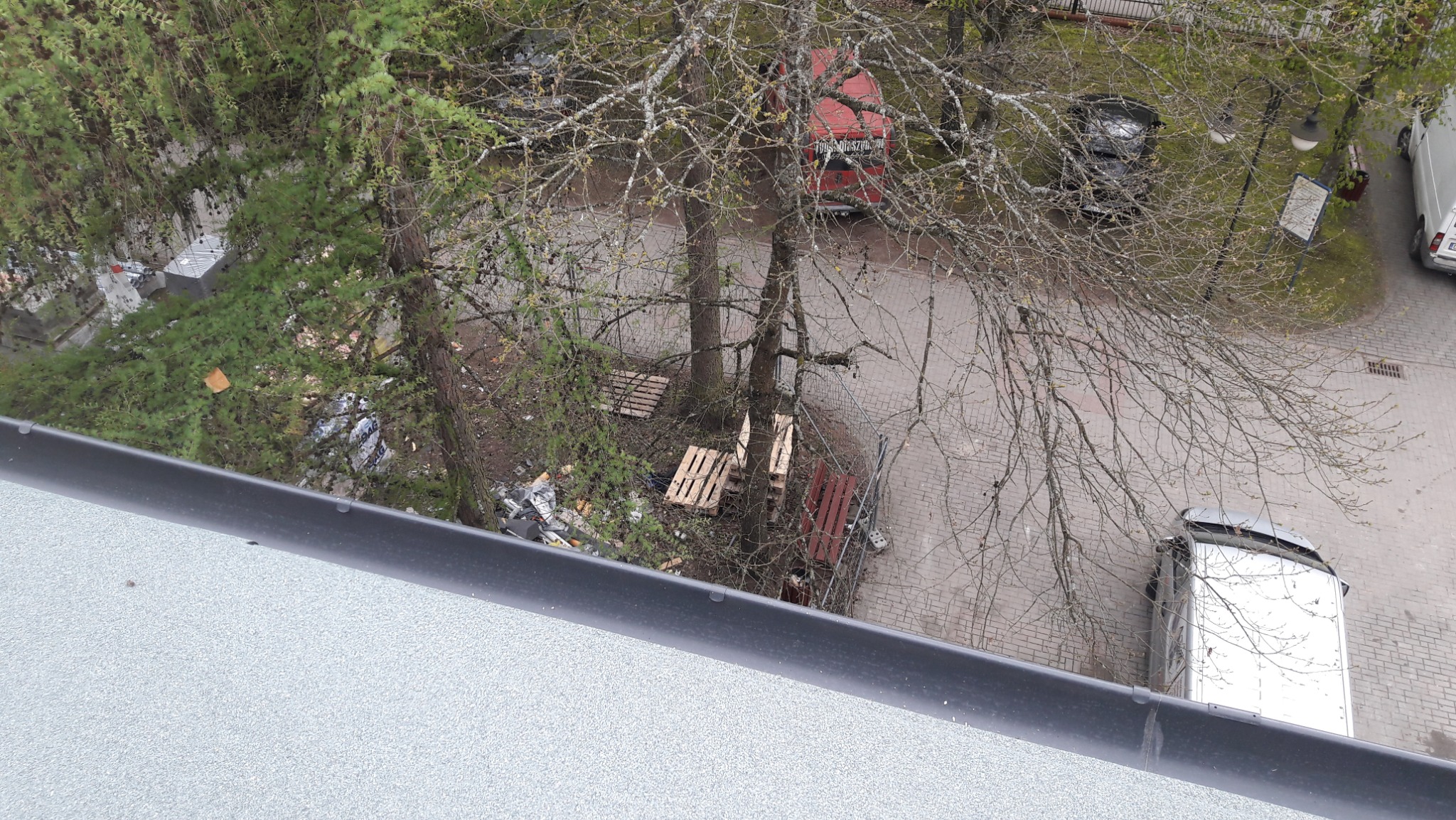 Widok z góry na podwórko z drzewami, zaparkowanymi samochodami i składowanymi paletami, widziany z dachu budynku z zamontowaną rynną.