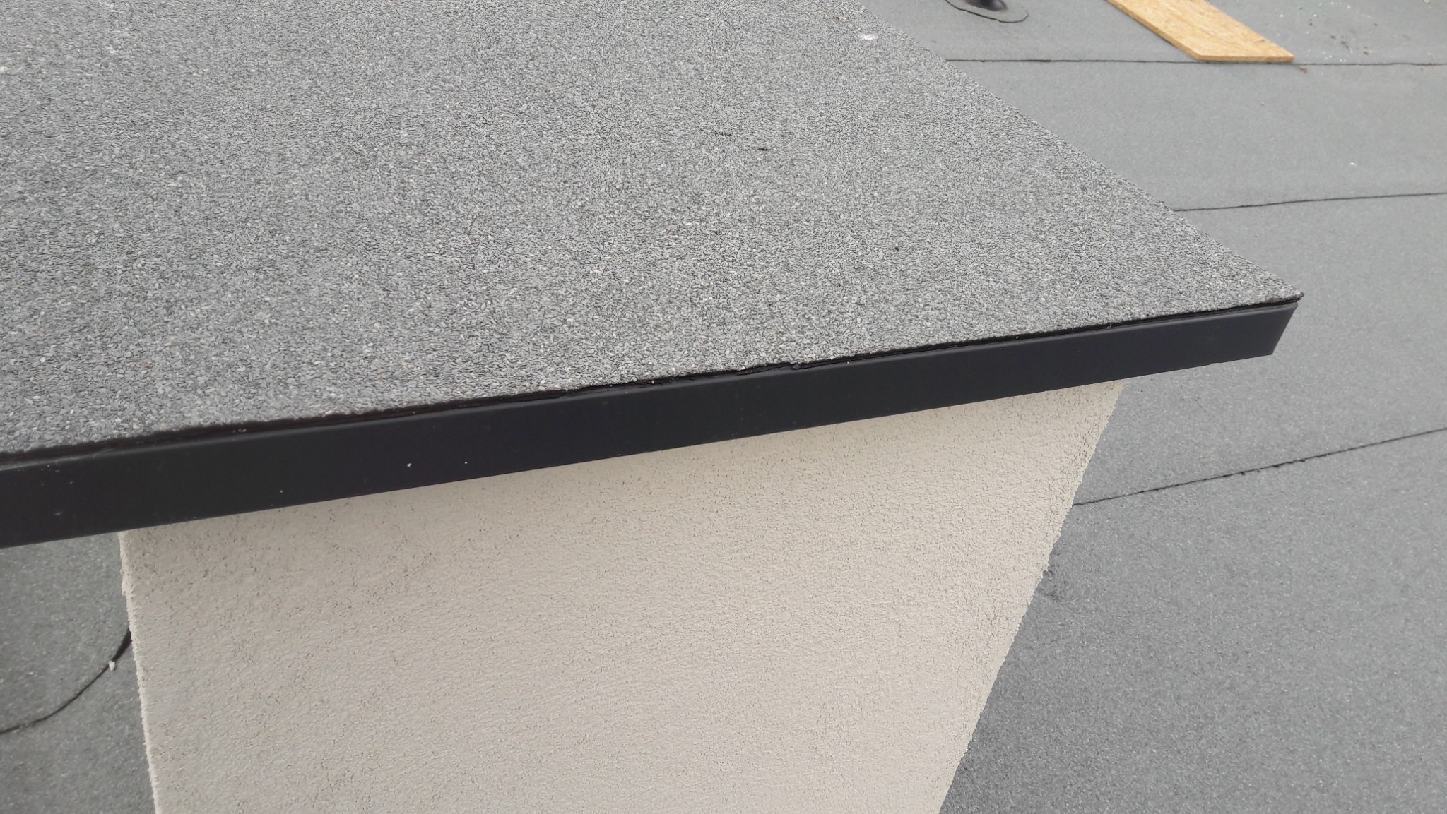 Detale dachu pokrytego papą z czarną listwą wykończeniową, fragment ściany szczytowej o jasnej, chropowatej fakturze.