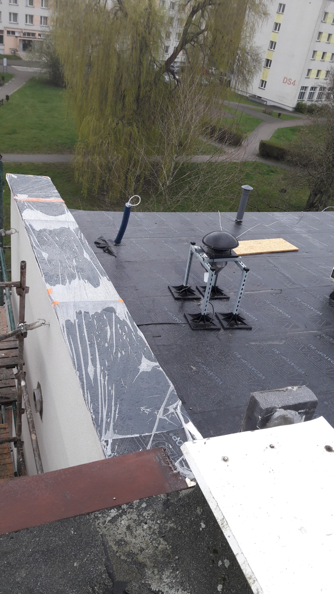 Prace na dachu bloku mieszkalnego, widoczna papa dachowa z nadrukiem, wentylator dachowy na metalowej konstrukcji, zabezpieczona ściana budynku, fragment rusztowania i widok na zieleń.