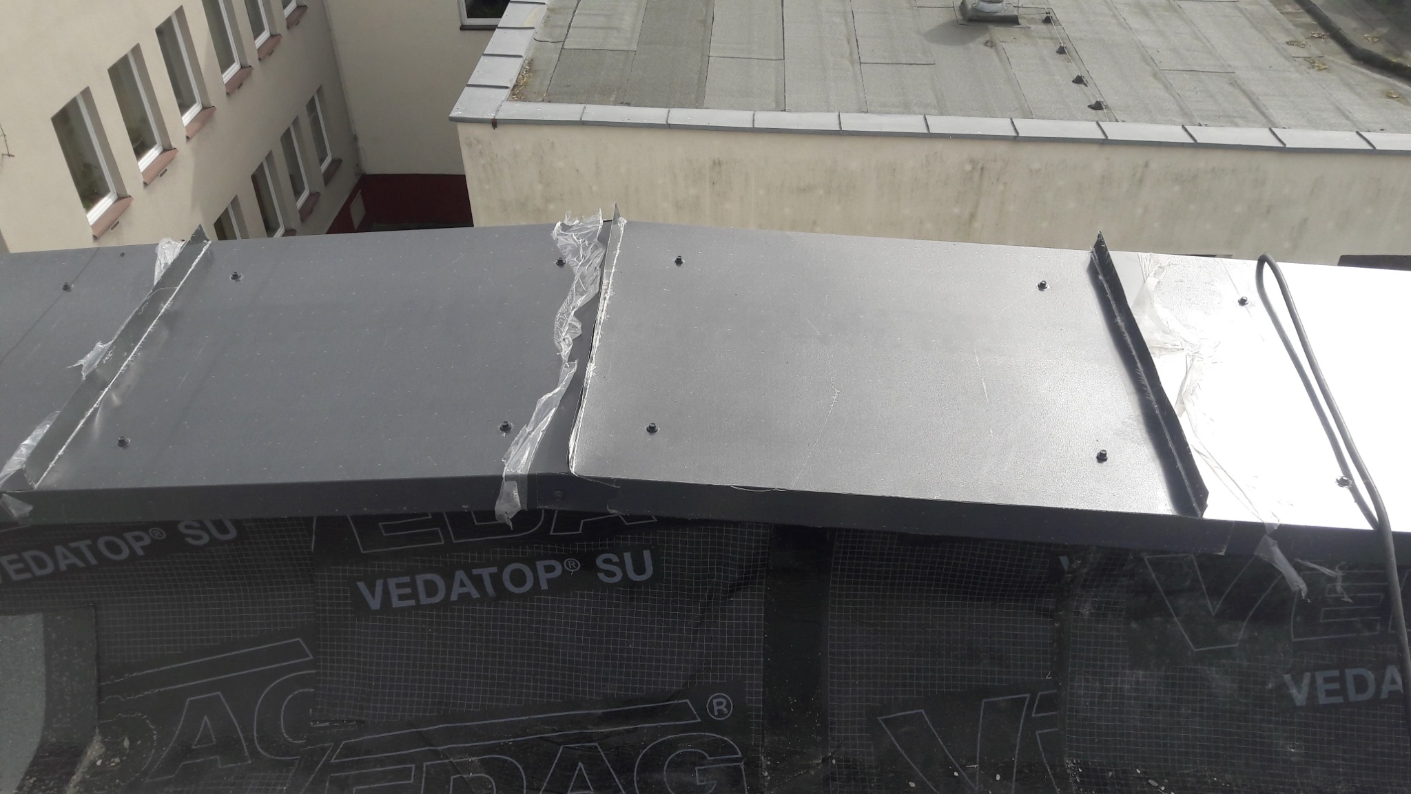 Szare panele blaszane mocowane do dachu pokrytego membraną hydroizolacyjną z widocznym logo producenta, w tle budynek z oknami.