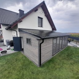 Kompleksowe usługi dekarsko ciesielskie - Przydomowa zabudowa tarasowa z drewna w szarym odcieniu, z czarnym dachem i rynnami, dobudowana do białego domu z ciemną dachówką, widok na trawnik i polską flagę w tle.