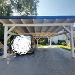 Nowoczesna wiata garażowa z jasnego drewna i ciemnym dachem, z zaparkowanym szarym vanem na wybrukowanym podjeździe, widok na zadbany ogród i dom w tle.