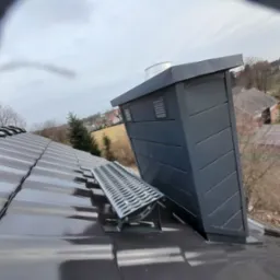 Nowoczesny komin wentylacyjny z metalową ławą kominiarską na błyszczącej, grafitowej dachówce ceramicznej, widok na osiedle domów jednorodzinnych w tle.