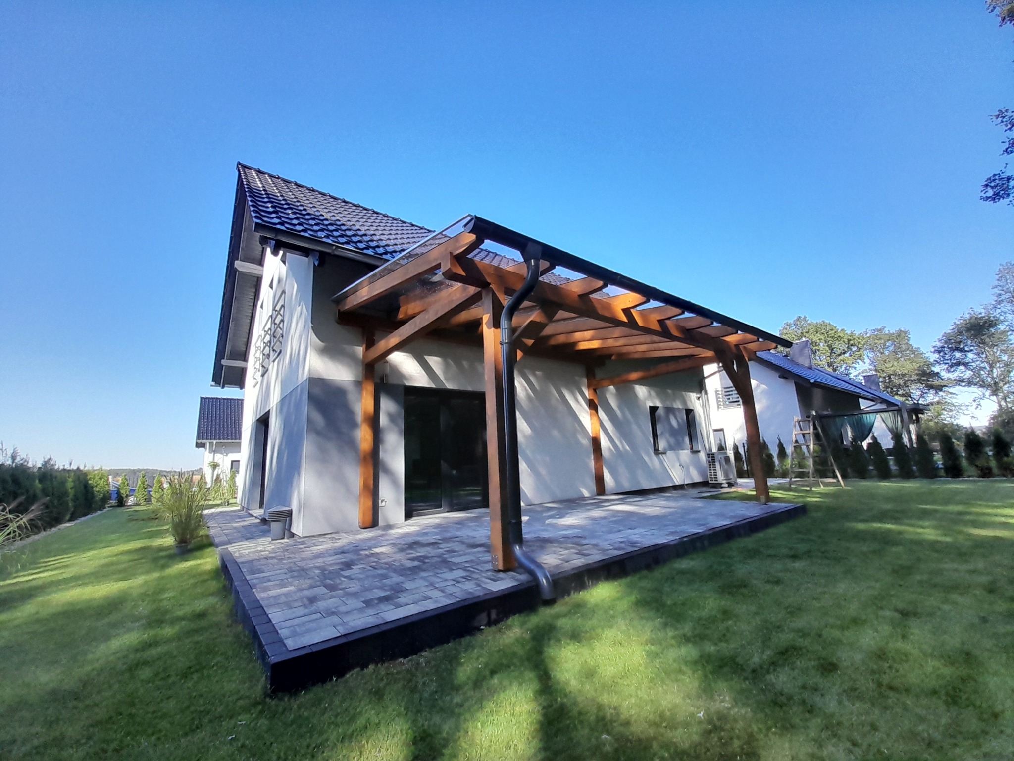 Drewniana pergola z przezroczystym zadaszeniem przylegająca do nowoczesnego domu w Nysie, widok z niskiej perspektywy na tle błękitnego nieba.