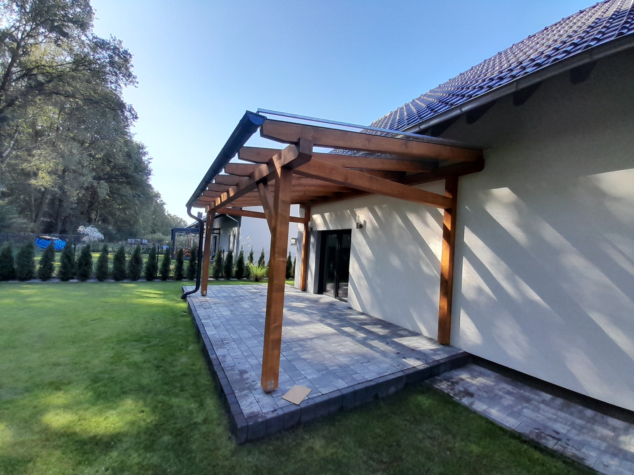 Nowoczesna konstrukcja z drewna z przezroczystym zadaszeniem, przylegająca do jasnej ściany budynku, z wybrukowanym tarasem i zadbanym trawnikiem. Widoczne ciemne rynny i rząd tuj w tle.