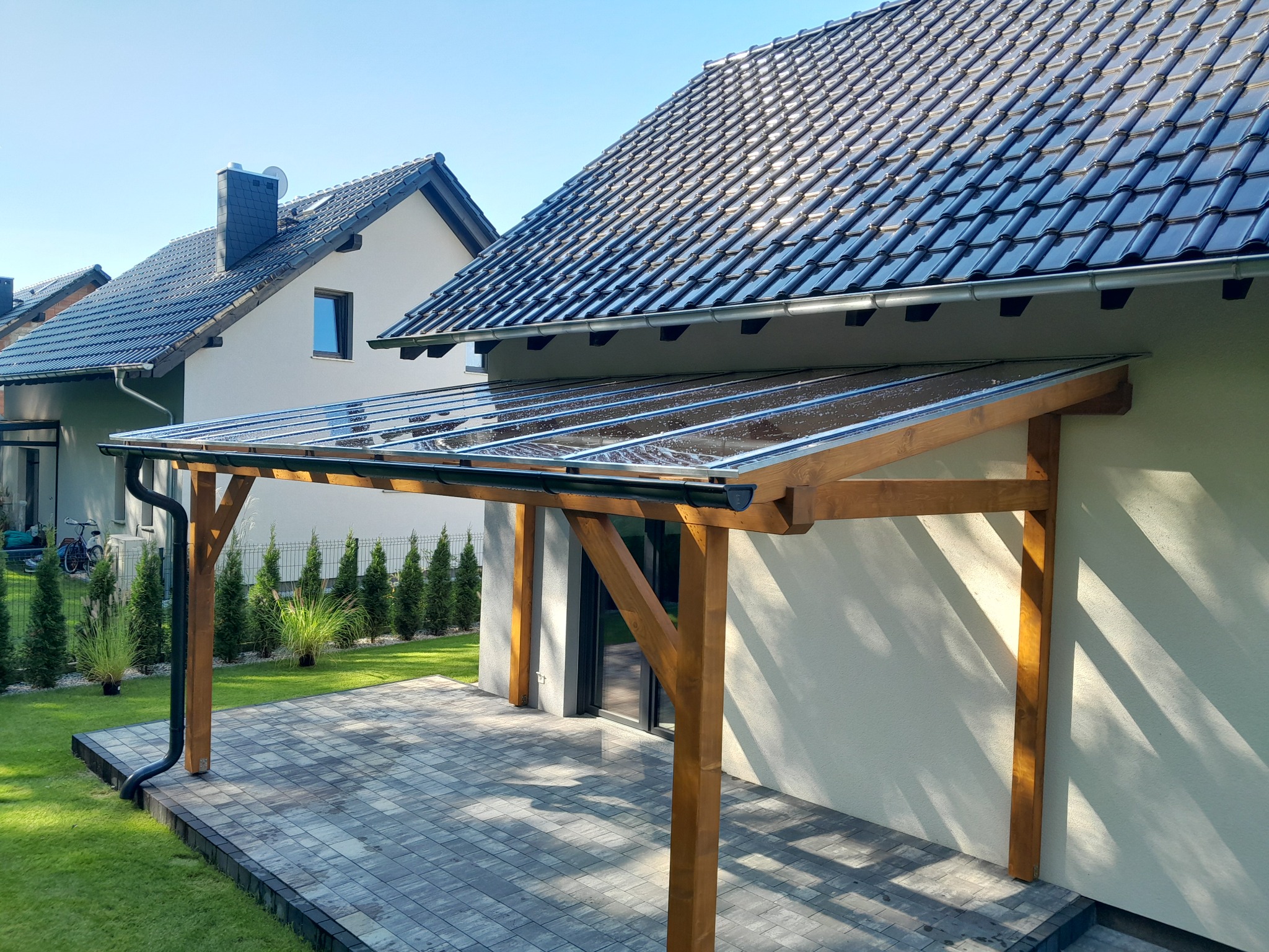 Zadaszenie tarasu z drewnianą konstrukcją i przezroczystym dachem, przylegające do domu z szarą elewacją i ciemną dachówką, widok na zadbany trawnik i sąsiedni budynek.