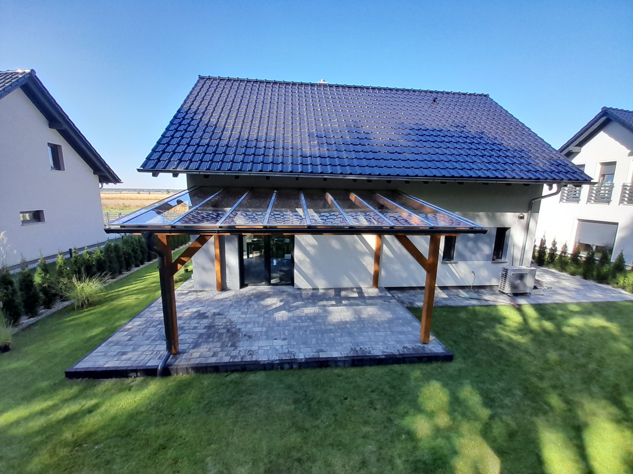 Zadaszony taras przydomowy z drewnianymi słupami i przezroczystym zadaszeniem, widok z góry na zadbany trawnik i kostkę brukową.