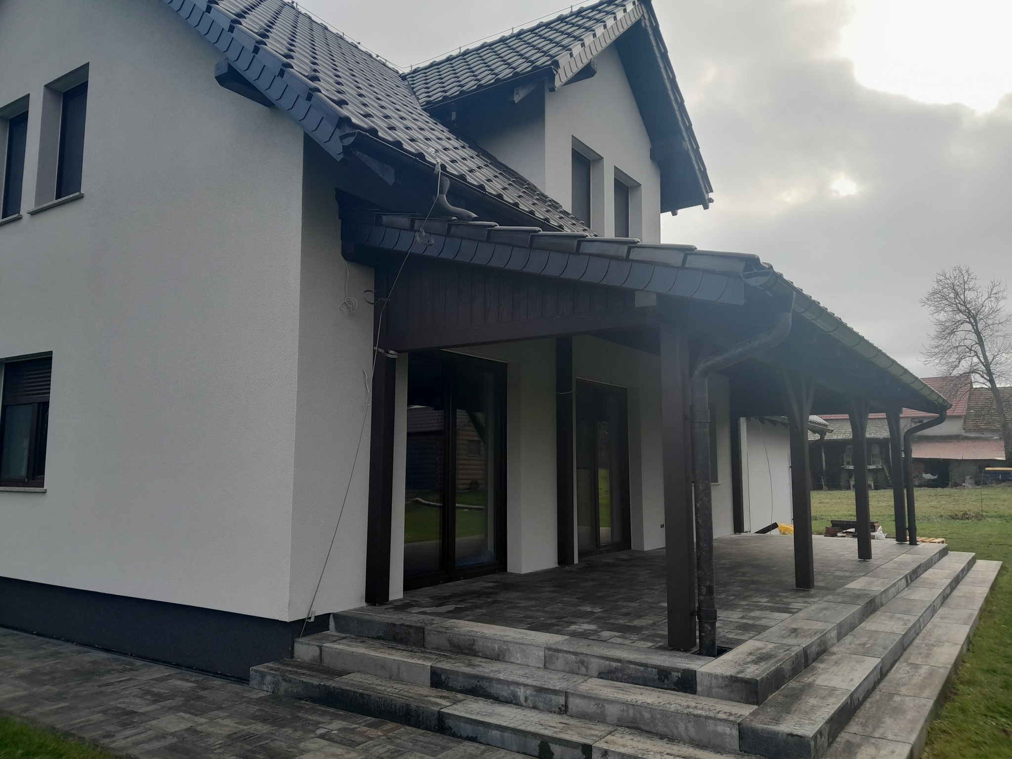 Nowoczesny dom z białymi ścianami i ciemnobrązową drewnianą konstrukcją zadaszenia tarasu, widoczne schody z szarej kostki brukowej i ciemna dachówka na dachu.