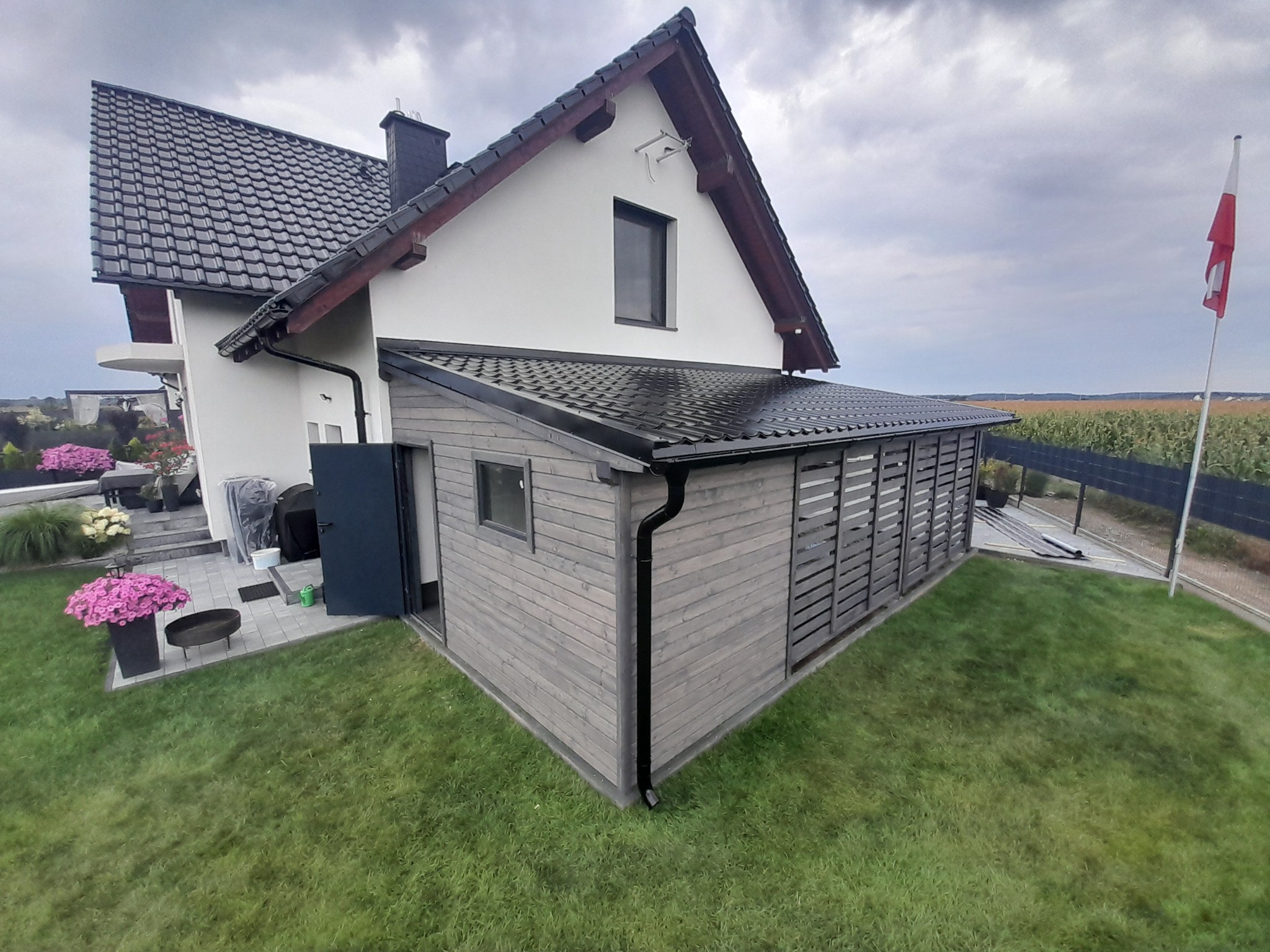 Przydomowa zabudowa tarasowa z drewna w szarym odcieniu, z czarnym dachem i rynnami, dobudowana do białego domu z ciemną dachówką, widok na trawnik i polską flagę w tle.