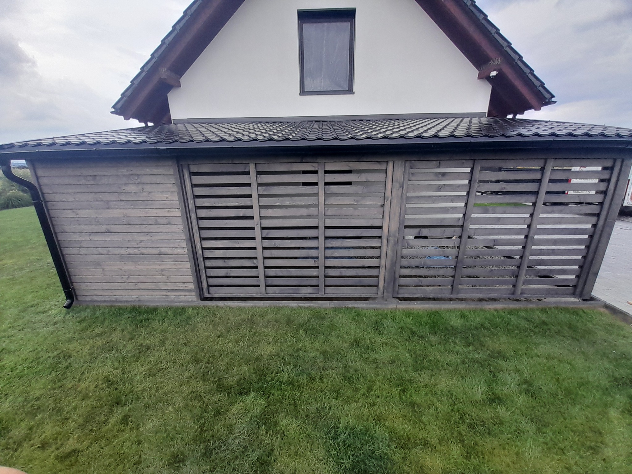Zadaszone miejsce postojowe z drewnianą konstrukcją i żaluzjowymi panelami, przylegające do domu z grafitową dachówką i rynną, otoczone trawnikiem.