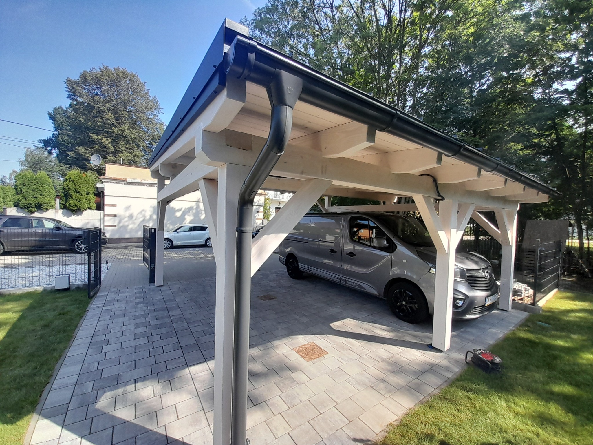 Nowoczesna, biała konstrukcja z drewna z czarnym dachem i rynnami, służąca jako zadaszenie dla samochodu na posesji z kostki brukowej, z widocznym sprzętem ogrodniczym na trawie.