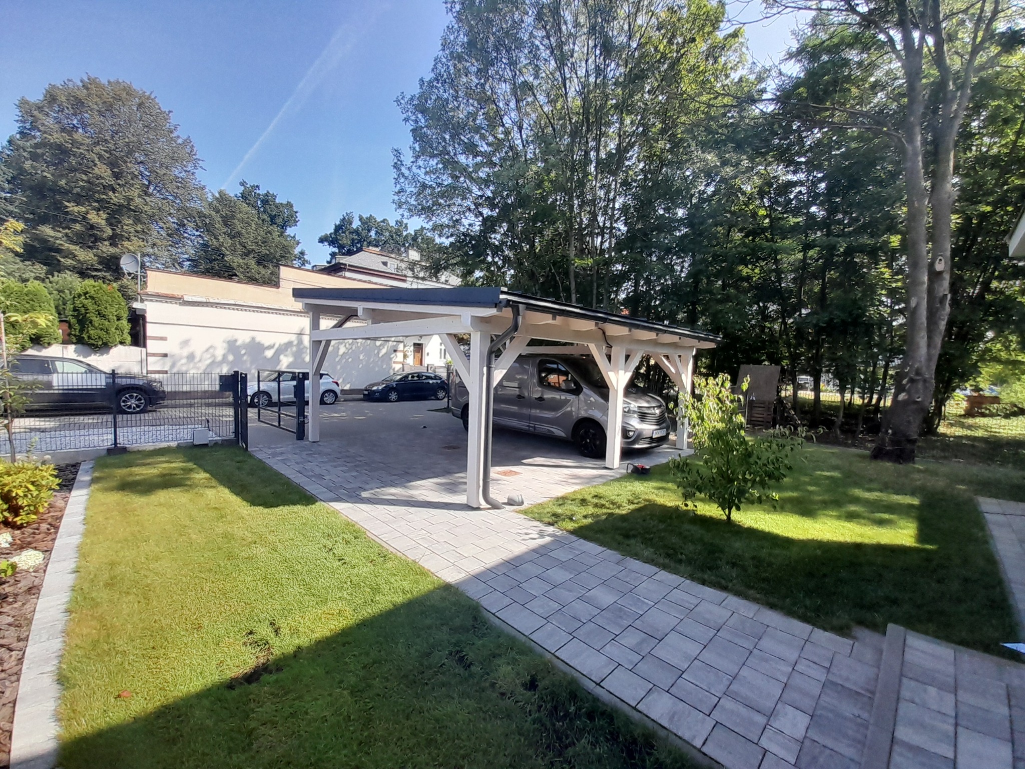 Nowoczesna, biała pergola drewniana z zadaszeniem, chroniąca zaparkowany samochód na posesji z zadbanym trawnikiem i kostką brukową.