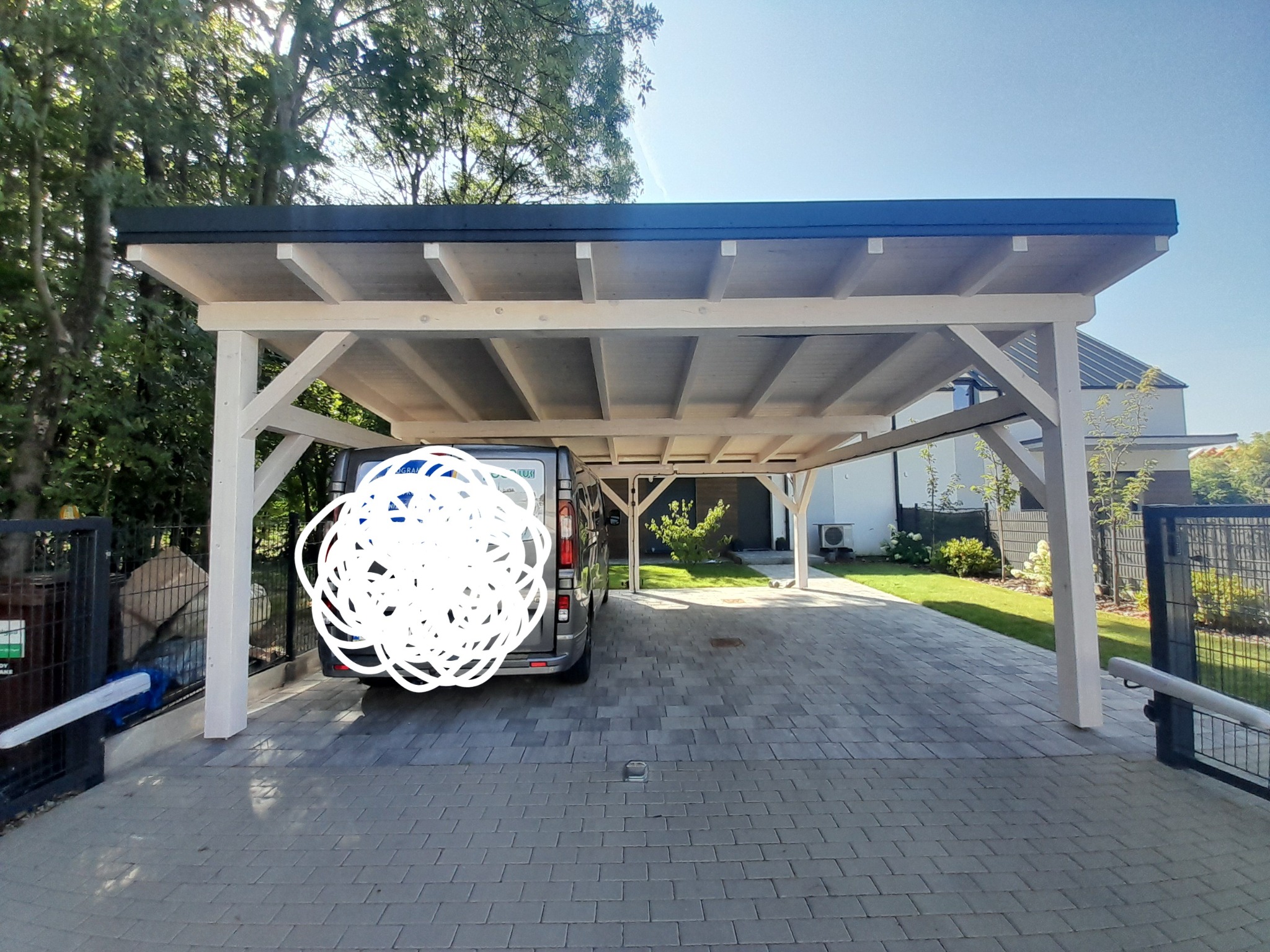 Nowoczesna wiata garażowa z jasnego drewna i ciemnym dachem, z zaparkowanym szarym vanem na wybrukowanym podjeździe, widok na zadbany ogród i dom w tle.