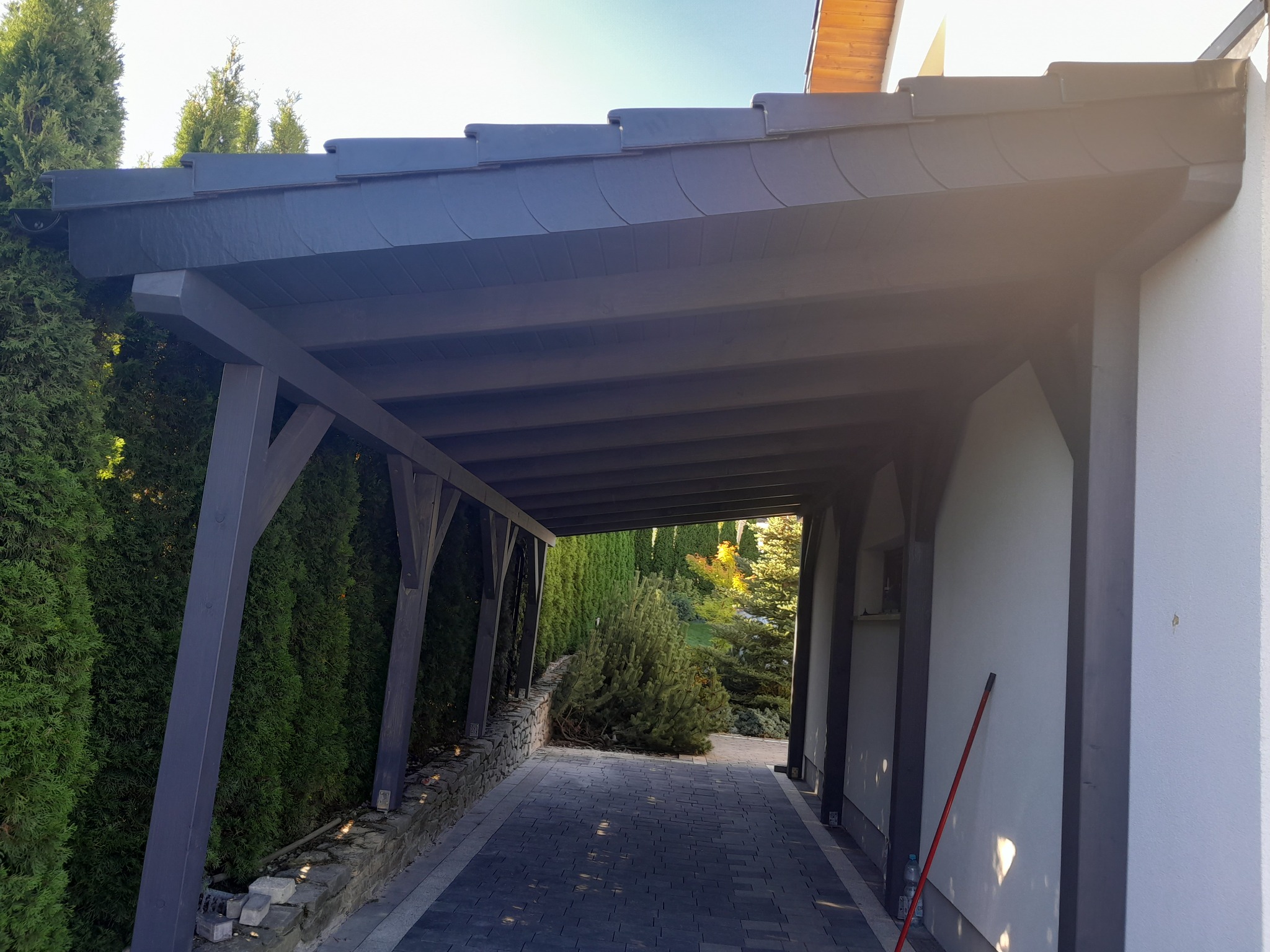 Zadaszone przejście z drewnianą konstrukcją pomalowaną na szaro, kryte ciemną dachówką, prowadzące przez posesję z widokiem na ogród.