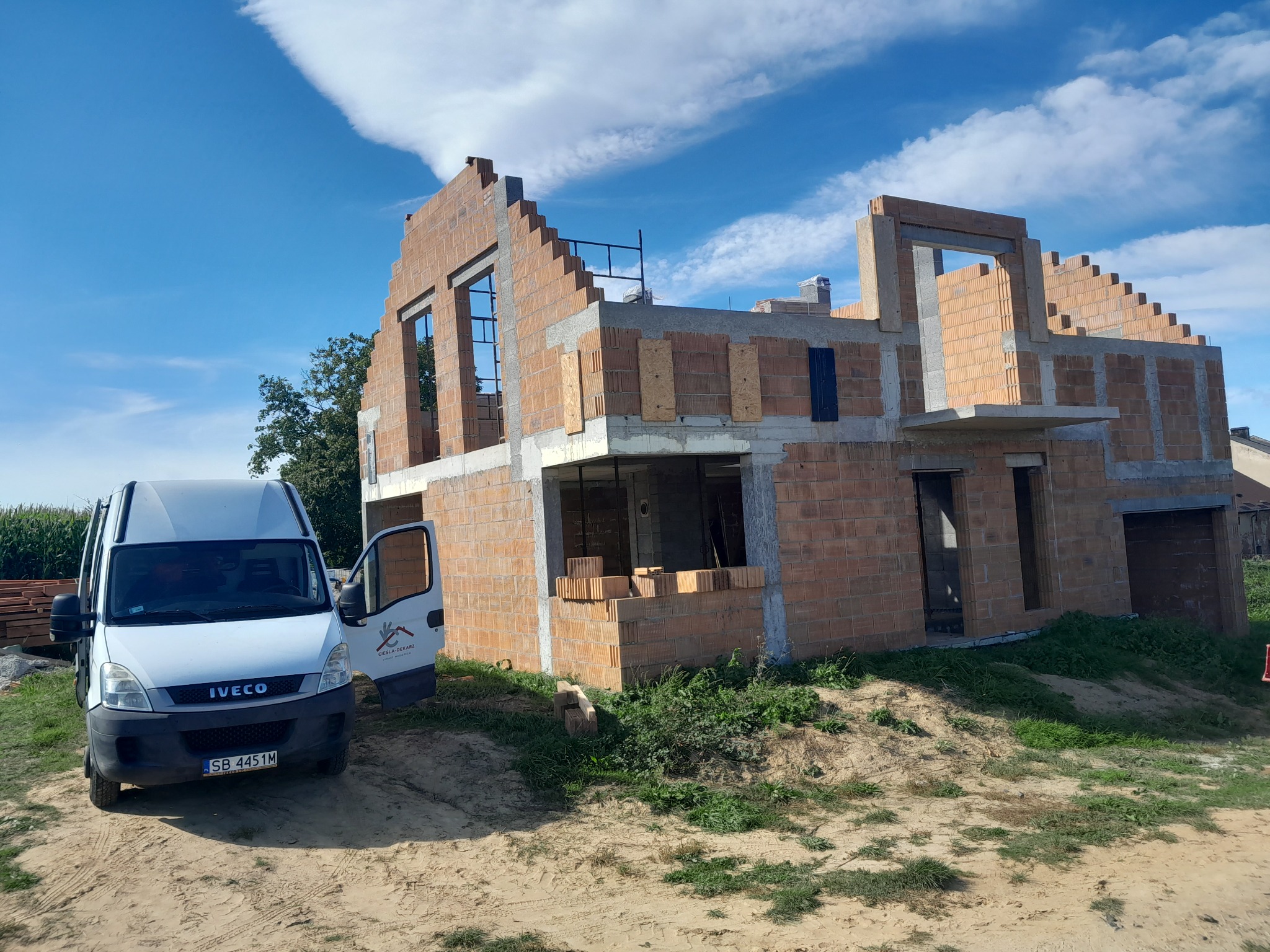 Wznoszenie dwukondygnacyjnego domu z czerwonej cegły, widoczny biały samochód dostawczy z otwartymi drzwiami i logo firmy budowlanej.