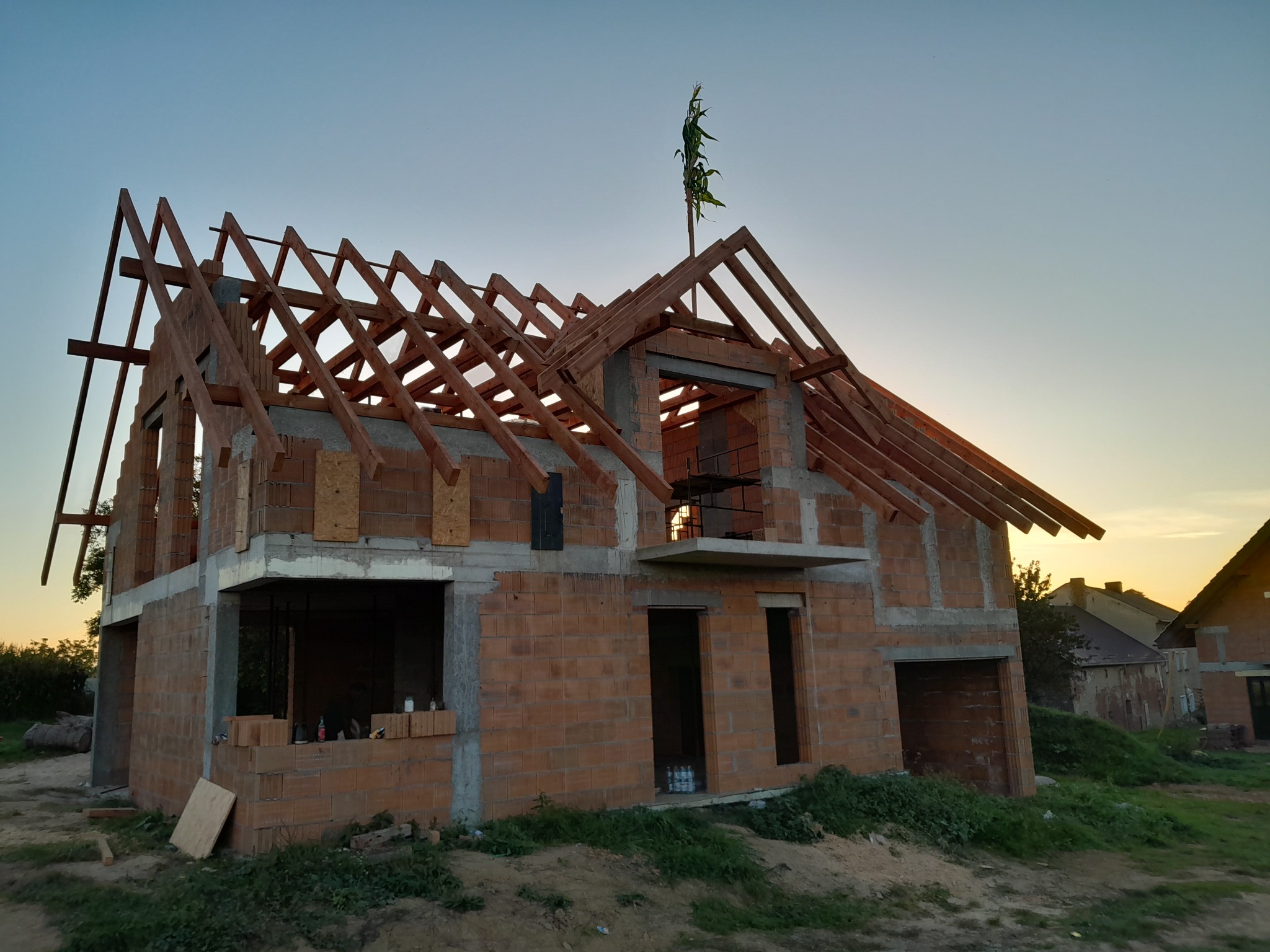 Surowy stan budynku z cegły z widoczną konstrukcją drewnianą dachu w trakcie budowy, z zieloną gałązką na szczycie, sfotografowany o zmierzchu.