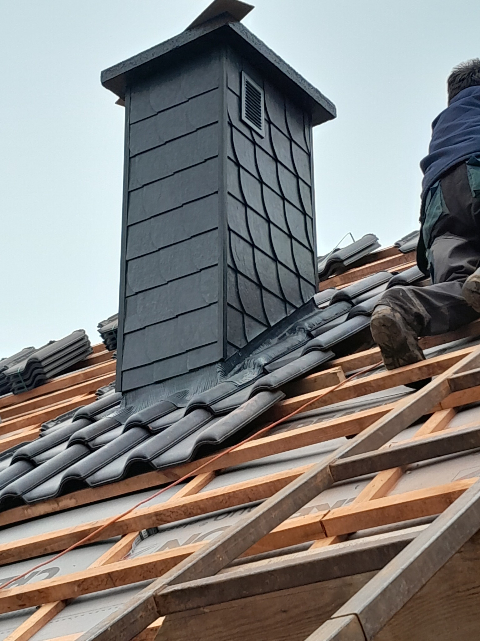 Murarz w roboczych butach klęczy na drewnianej konstrukcji dachu z czarną dachówką, obok komin wykończony łupkiem, w tle pochmurne niebo.