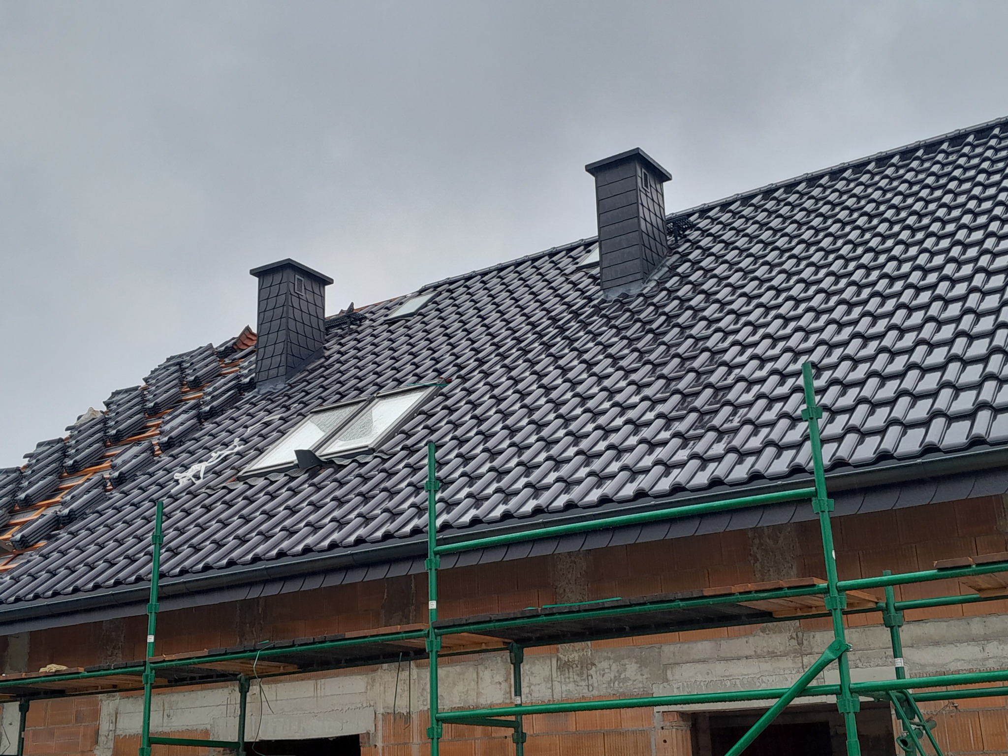Częściowo pokryty ciemną dachówką dach z oknami połaciowymi i kominami, widoczne rusztowanie i mur budynku w trakcie prac.