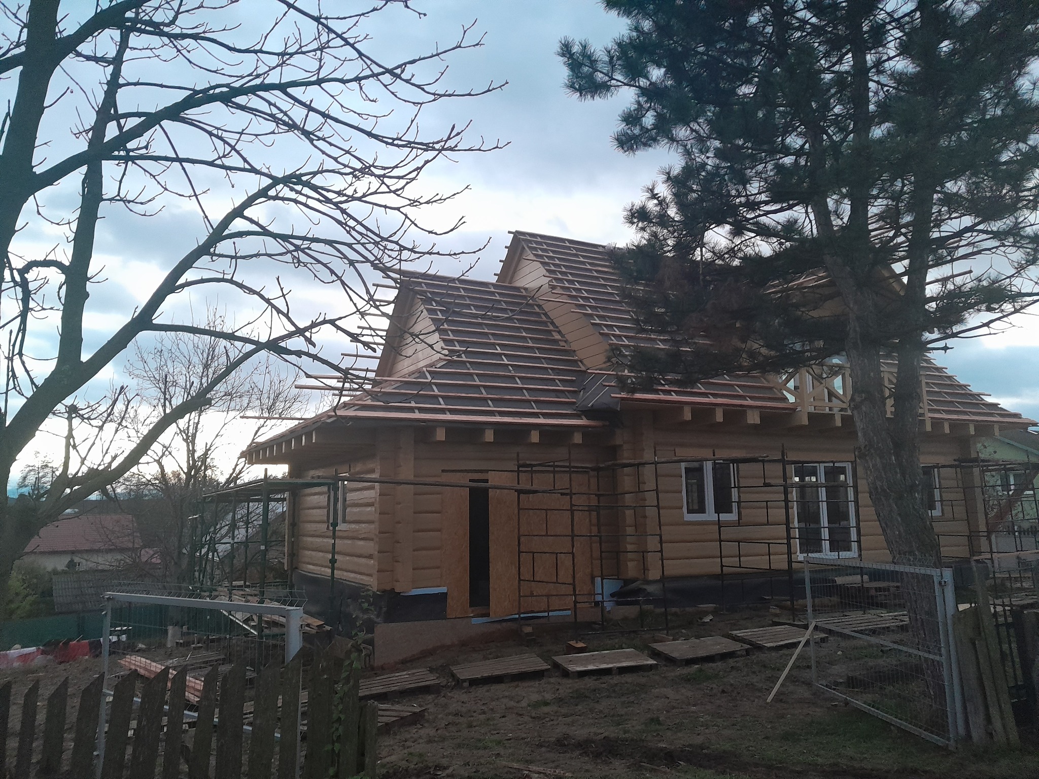 Budowa domu z bali w trakcie wznoszenia dachu, widoczne rusztowania i elementy konstrukcyjne, drzewa w tle.