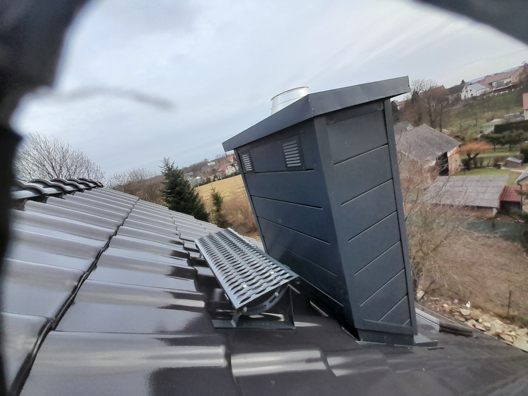 Nowoczesny komin wentylacyjny z metalową ławą kominiarską na błyszczącej, grafitowej dachówce ceramicznej, widok na osiedle domów jednorodzinnych w tle.