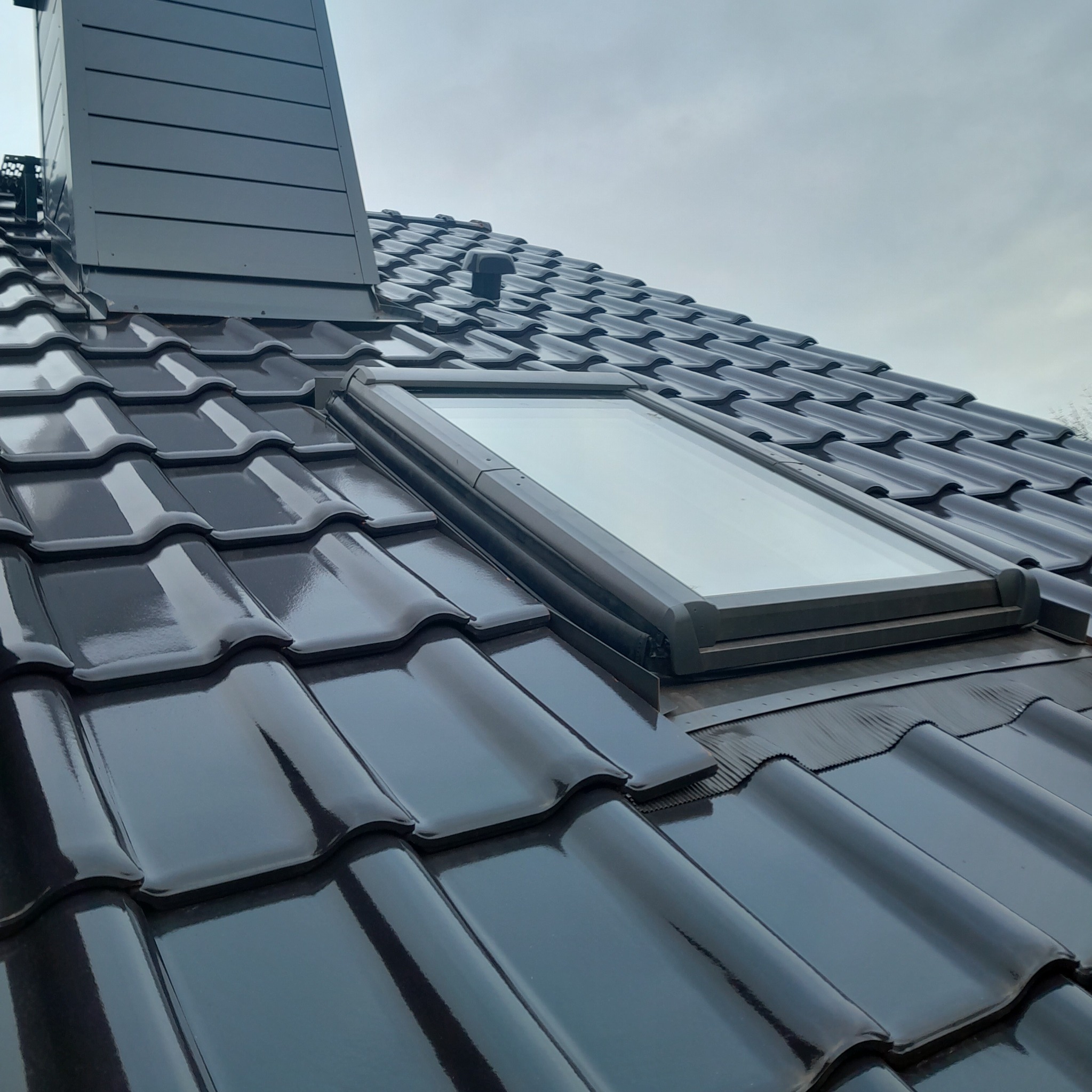 Błyszcząca, grafitowa dachówka ceramiczna z oknem dachowym i kominem wentylacyjnym, widok z dołu pod kątem na pochmurnym niebie.
