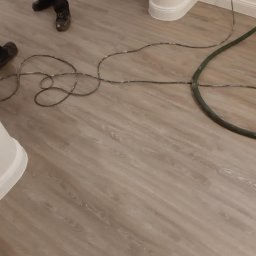 Konrad Zelewski - Jasne panele podłogowe z widocznymi kablami i wężem, fragment ściany z listwą przypodłogową i gniazdkiem elektrycznym, buty robocze w tle.