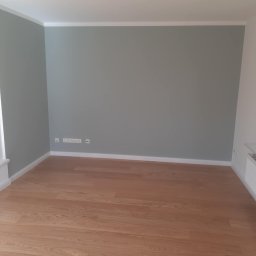 Konrad Zelewski - Pomalowane na szaro wnętrze pokoju z drewnianą podłogą, białymi listwami przypodłogowymi i kontaktami elektrycznymi na ścianie.