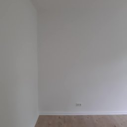 Konrad Zelewski - Pusty, świeżo wyremontowany pokój z jasnymi ścianami, drewnianą podłogą i gniazdkiem elektrycznym przy listwie przypodłogowej.