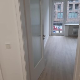 Konrad Zelewski - Otwarta biała, przeszklona drzwi wewnętrzne, widok na jasne panele podłogowe w pustym pokoju z oknem na budynek mieszkalny.