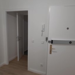 Konrad Zelewski - Wnętrze mieszkania z dwiema białymi drzwiami: jedne wejściowe z szarą listwą antypaniczną i domofonem, drugie otwarte prowadzące do innego pomieszczenia, jasna podłoga.