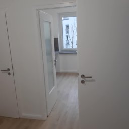 Konrad Zelewski - Wnętrze mieszkania z widokiem na otwarte białe drzwi z przeszkleniem, ukazujące podłogę z paneli i okno z widokiem na budynek.