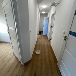 Konrad Zelewski - Korytarz w trakcie remontu z świeżo pomalowanymi ścianami na biało, drewnopodobną podłogą, otwartymi białymi drzwiami z przeszkleniami i białymi drzwiami z niebieskim paskiem po prawej stronie...