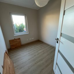 Konrad Zelewski - Pokój z panelami podłogowymi, widoczne fragmenty drzwi z srebrnymi wstawkami, okno z widokiem na zieleń i osobę, oraz lampa w stylu japońskim pod sufitem. Na podłodze leżą panele przed montażem.