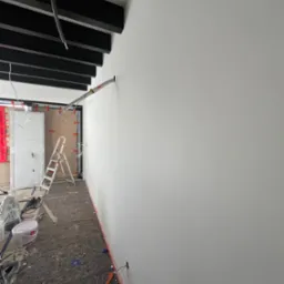 Wnętrze w trakcie remontu, ściana pomalowana na biało, widoczne przewody, drabina, wiaderka z farbą i zabezpieczona podłoga.