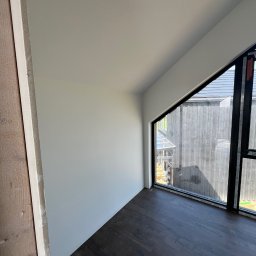 Konrad Zelewski - Wnętrze pokoju z ukośną ścianą i oknem o nieregularnym kształcie, widoczne świeżo położone panele podłogowe w ciemnym kolorze, ściany pomalowane na biało, na jednej ze ścian widoczny surowy...