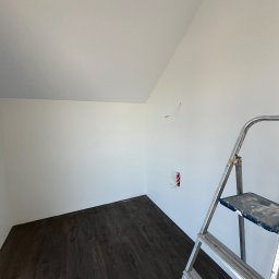 Konrad Zelewski - Poddasze z ciemną podłogą z paneli, białe ściany po świeżym malowaniu, aluminiowa drabina z plamami farby stoi w rogu, widoczne wystające przewody elektryczne.