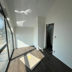 Konrad Zelewski - Jasne wnętrze w stanie deweloperskim z dużym oknem, panelami na podłodze i świeżo pomalowanymi ścianami na biało. Na podłodze leży karton ochronny z żółtą plamą. W głębi widoczne otwarte drzwi...