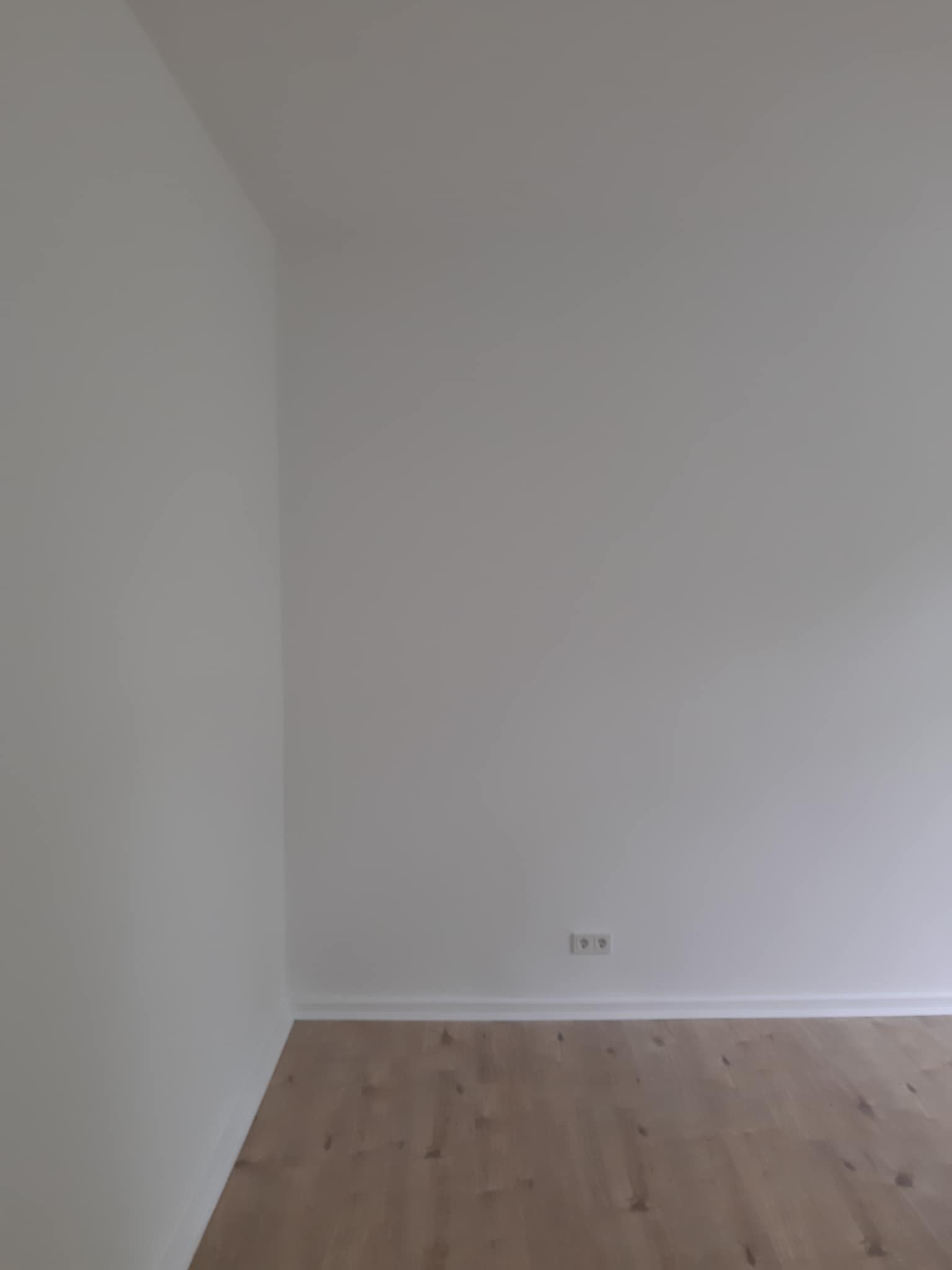 Pusty, świeżo wyremontowany pokój z jasnymi ścianami, drewnianą podłogą i gniazdkiem elektrycznym przy listwie przypodłogowej.