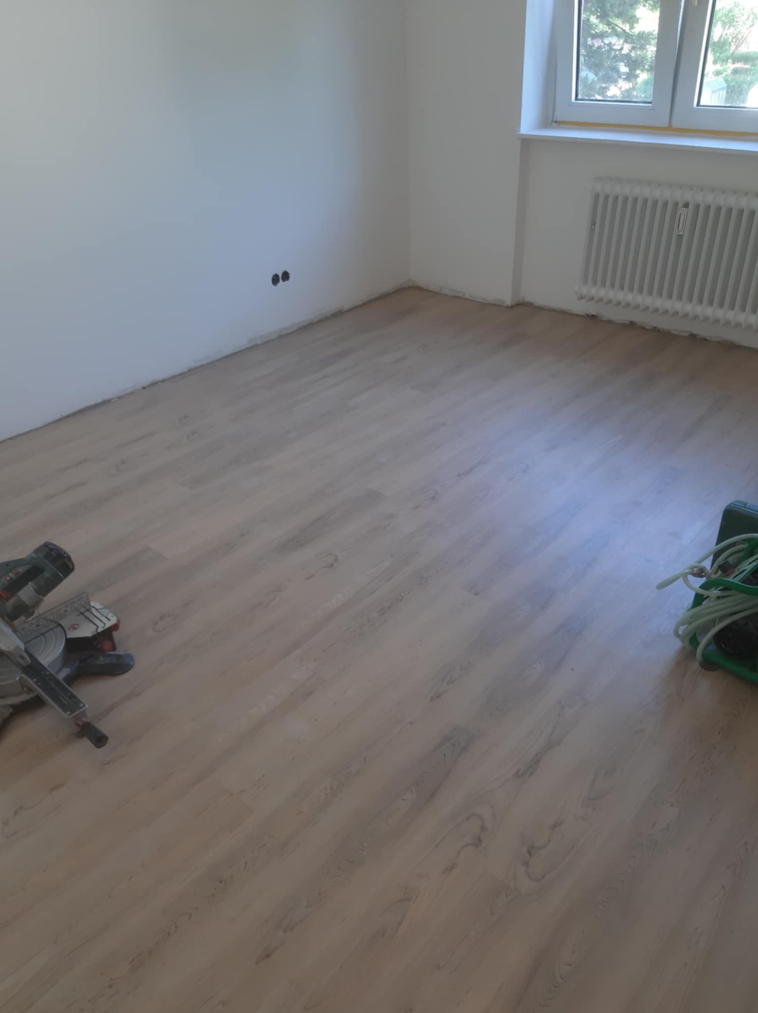Układanie paneli podłogowych w pokoju, widoczna ukośnica i listwa przypodłogowa w trakcie montażu, grzejnik pod oknem.