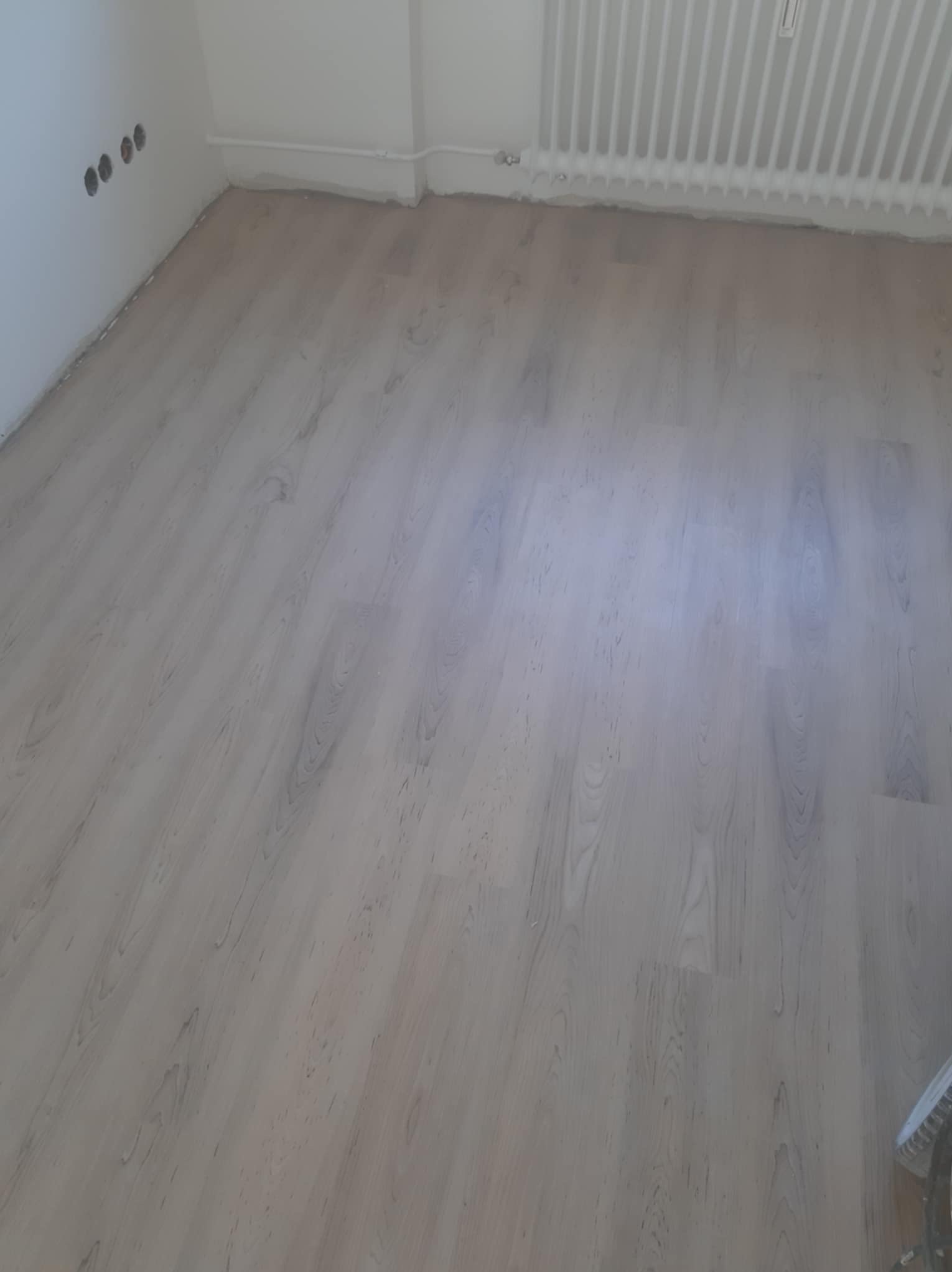 Jasne panele podłogowe z widocznym wzorem drewna, ułożone w pomieszczeniu z białymi ścianami i grzejnikiem, widoczne otwory na gniazdka elektryczne w ścianie.