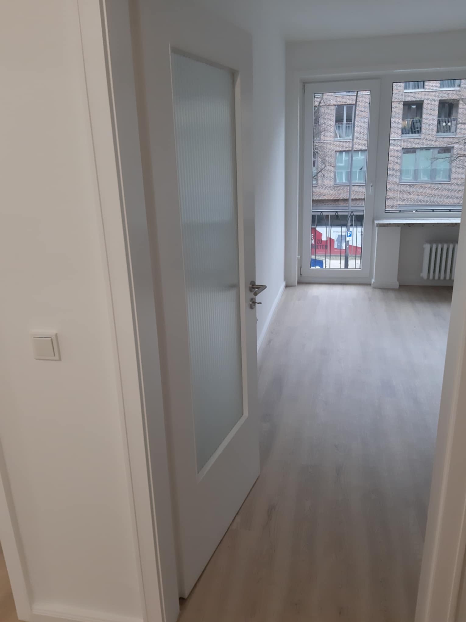 Otwarta biała, przeszklona drzwi wewnętrzne, widok na jasne panele podłogowe w pustym pokoju z oknem na budynek mieszkalny.