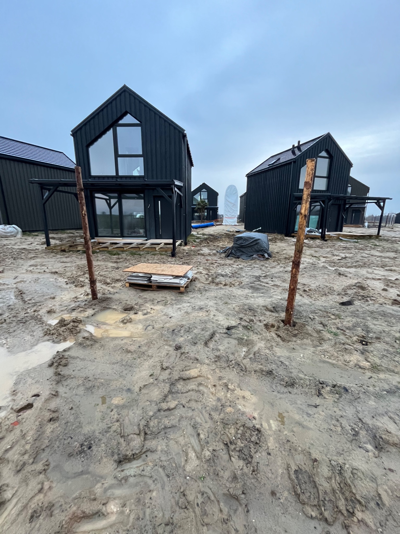 Wizualizacja osiedla domów w stylu nowoczesnej stodoły z czarną elewacją, w trakcie prac wykończeniowych na zewnątrz, widoczne drewniane pale, palety z płytami i błotniste podłoże.