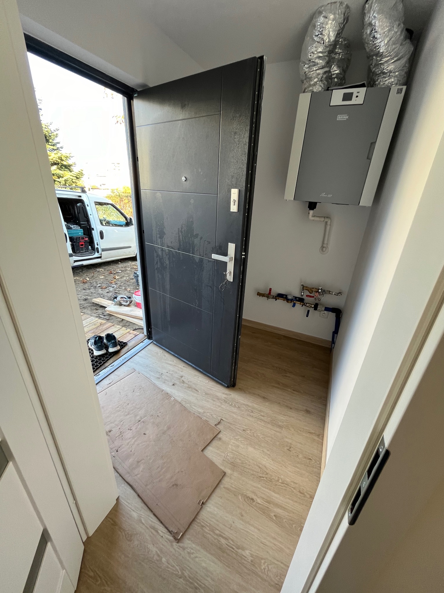 Nowoczesne drzwi wejściowe w trakcie montażu, widoczny sprzęt wentylacyjny na ścianie, karton zabezpieczający podłogę i narzędzia w tle.