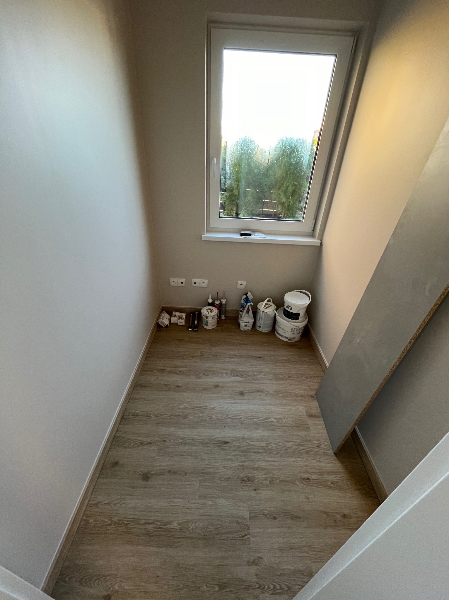 Wnętrze pomieszczenia z beżowymi ścianami, drewnopodobną podłogą i oknem, widoczne puszki z farbą, narzędzia malarskie oraz płyta meblowa oparta o ścianę.