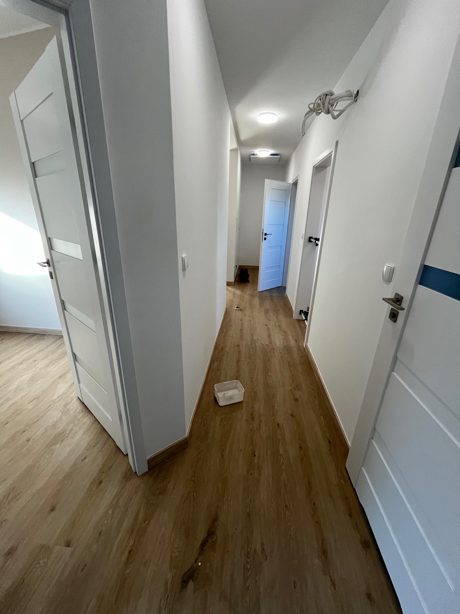 Korytarz w trakcie remontu z świeżo pomalowanymi ścianami na biało, drewnopodobną podłogą, otwartymi białymi drzwiami z przeszkleniami i białymi drzwiami z niebieskim paskiem po prawej stronie...