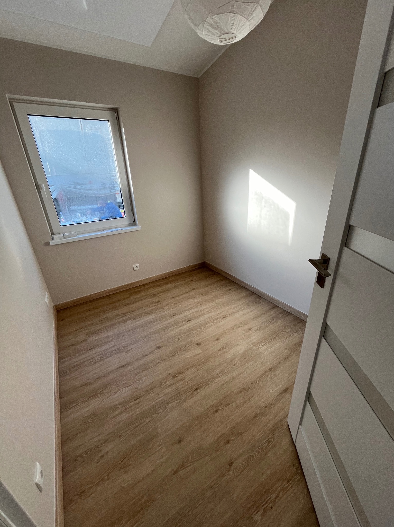 Pusty pokój po remoncie z panelami podłogowymi w odcieniach drewna, świeżo pomalowanymi ścianami na beżowo, białymi drzwiami z ciemną klamką i oknem z widokiem na zewnątrz.