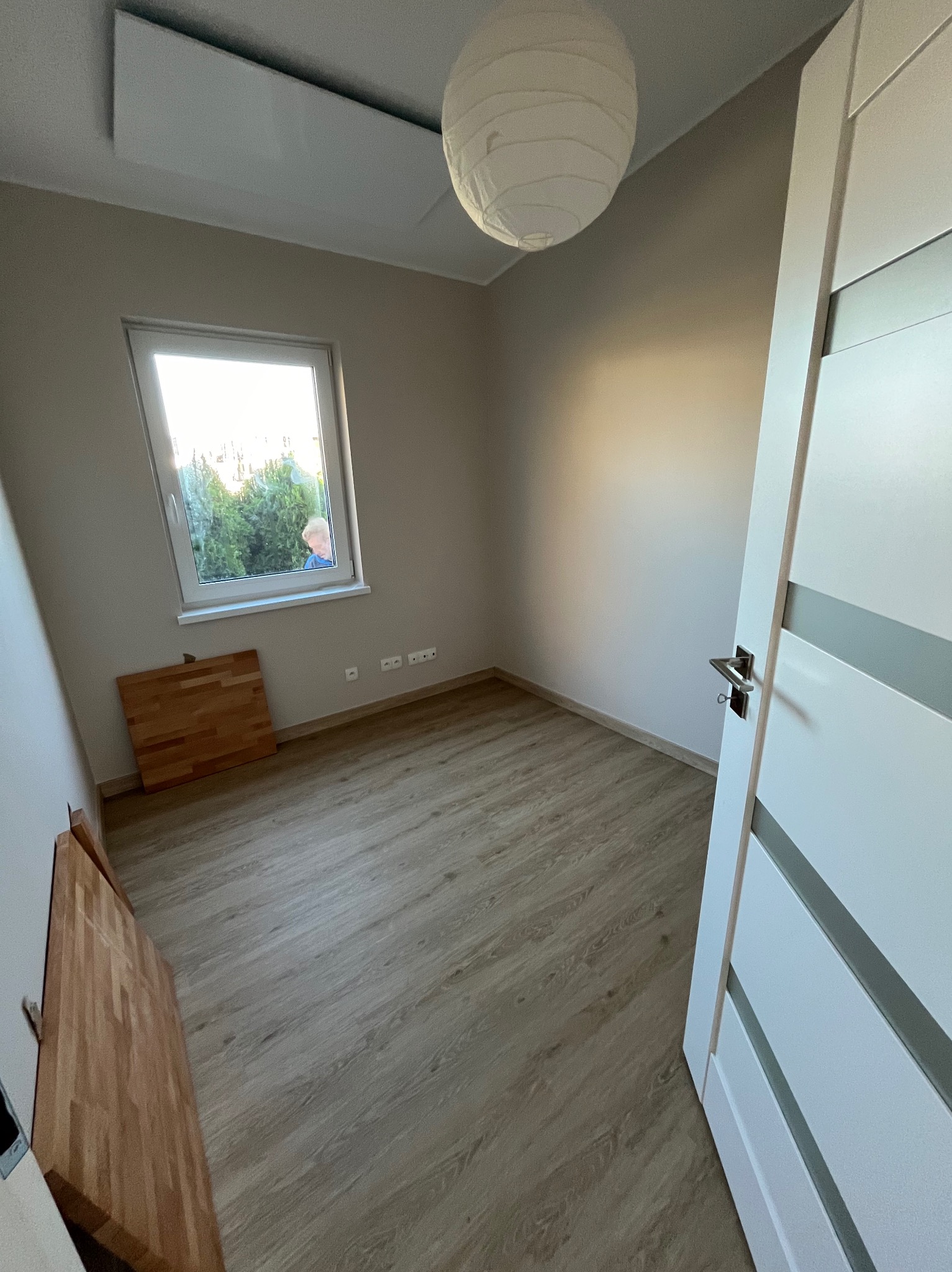 Pokój z panelami podłogowymi, widoczne fragmenty drzwi z srebrnymi wstawkami, okno z widokiem na zieleń i osobę, oraz lampa w stylu japońskim pod sufitem. Na podłodze leżą panele przed montażem.